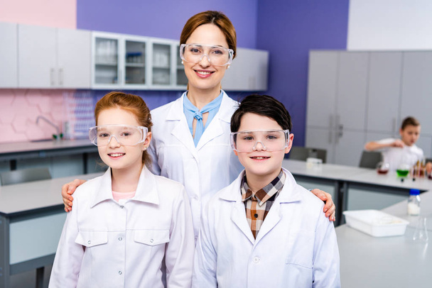 Uśmiechnięty nauczyciel w białej sierści i okulary ochronne obejmując uczniów podczas lekcji chemii - Zdjęcie, obraz