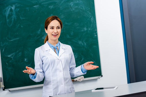Smiling chemistry teacher in white coat explaining lesson in front of blackboard - Photo, image