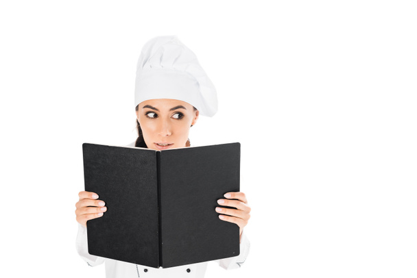 Nieuwsgierig chef-kok in uniform holding Zwartboek geïsoleerd op wit - Foto, afbeelding