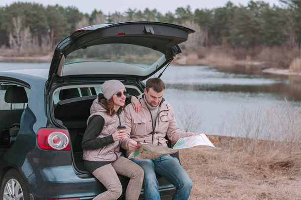 Young man and woman sitting in open trunk of car near the river and watching road map. - Photo, Image
