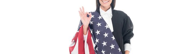 Πανοραμικό στιγμιότυπο της χαμογελώντας δικαστής κρατώντας την αμερικανική σημαία και δείχνει εντάξει σημάδι που απομονώνονται σε λευκό - Φωτογραφία, εικόνα