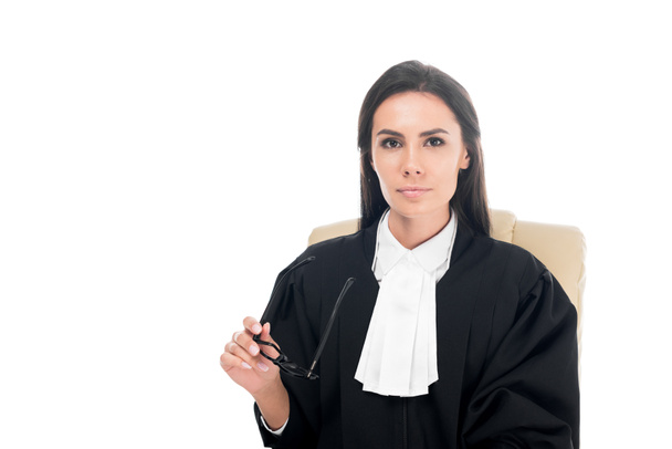 Beautiful judge in judicial robe sitting in armchair and holding glasses isolated on white - Photo, Image