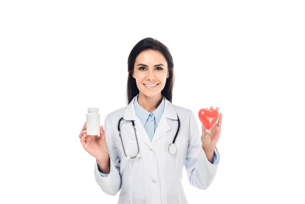 Doctor en bata blanca con estetoscopio sosteniendo píldoras y corazón de plástico aislado en blanco - Foto, imagen