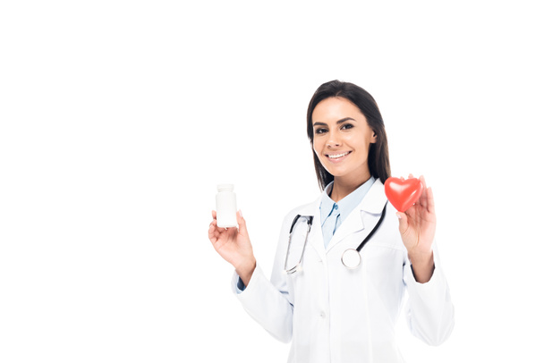 Doutor em casaco branco com estetoscópio segurando pílulas e coração de plástico isolado em branco - Foto, Imagem