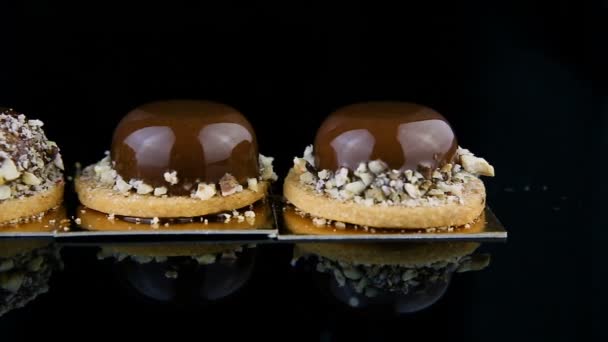 Πανόραμα από τρία Γαλλικά μίνι στρογγυλή μους γλυκά ζύμης καλυμμένο με γλάσο σοκολάτας και διακοσμημένα με θρυμματισμένα καρύδια που σερβίρεται σε φόντο μαύρο καθρέφτη - Πλάνα, βίντεο