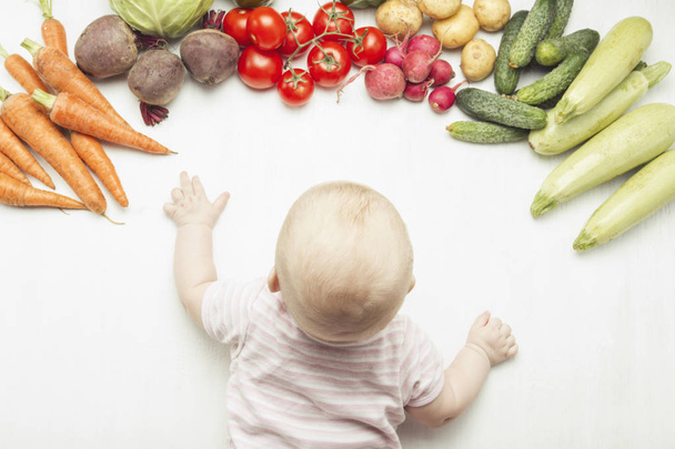 Little baby looks at fresh organic vegetables on a light wooden background. Care for health. Choice of the first food. Healthy baby food. Flat lay, top view - Фото, зображення