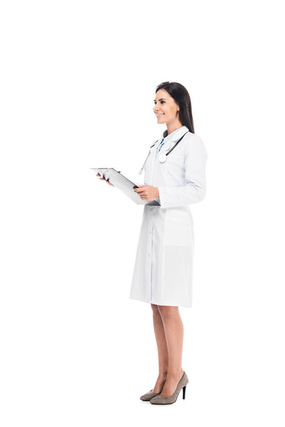 Full length of smiling doctor in white coat holding clipboard isolated on white - 写真・画像