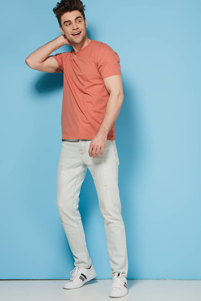 handsome and brunette man in t-shirt looking away and smiling on blue background  - Foto, afbeelding