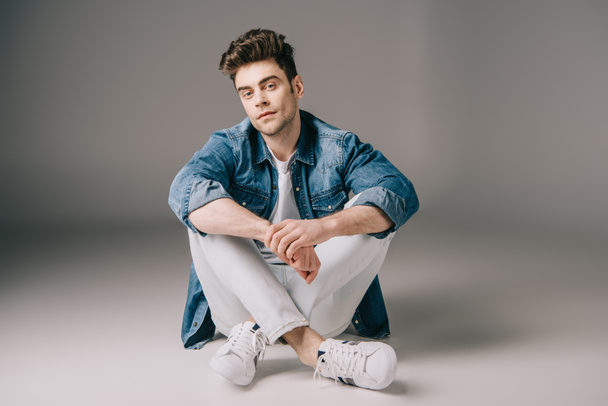 handsome man in denim shirt and jeans sitting on floor and looking at camera  - Zdjęcie, obraz