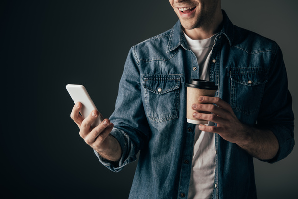 cropped view of man in denim shirt holding paper cup and using smartphone isolated on black - Foto, afbeelding