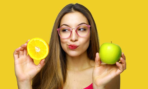 Wątpiący kobieta trzyma pół pomarańczowe i zielone jabłko i patrząc - Zdjęcie, obraz