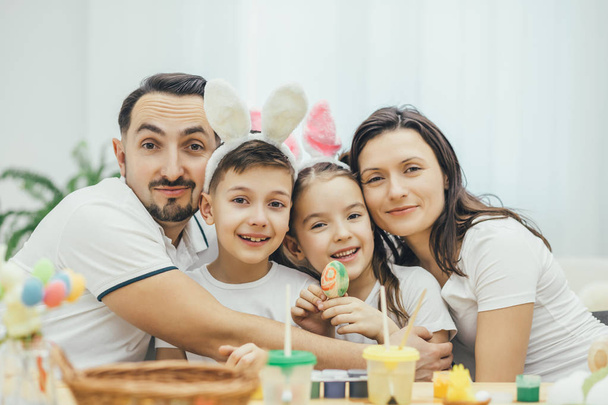 Happy family sitting at the table full og easter supplies, hugging and smiling. Children in funny bunny ears. Cute girl with pigtails is holding a colored egg. - Zdjęcie, obraz