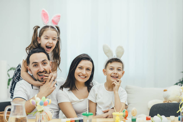 Happy family sitting at the table full of easter supplies, daughter is hugging her father. Father pretends to bite her finger. Little son is sitting on his mothers knees. Everyone is smiling. - Foto, Imagen