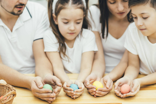Inspired family in white t-shirts sitting together at the table, holding colored easter eggs in their hands, looking at them excitedly, choosing which egg is the best. - Foto, Bild