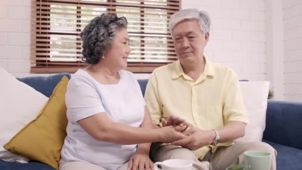 Asian elderly couple holding their hands while taking together in living room, couple feeling happy share and support each other lying on sofa at home. Lifestyle Senior family at home concept. - Footage, Video
