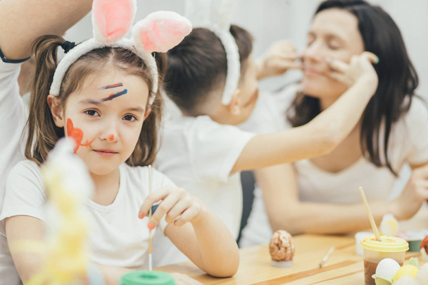 Family are ready for Easter. Family playing around. Lovely girl in bunny ears and with colored face on the forefront. Mother and son playing around on the blurred background. - Фото, изображение