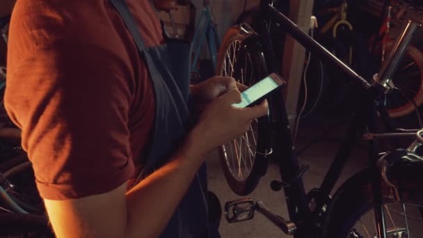 θέμα μικρών επιχειρήσεων επισκευής ποδηλάτου. Καυκάσιος μελαχρινή νεαρός φορώντας προστατευτικά γυαλιά, γάντια και fartukhe χρησιμοποιεί τεχνολογία κινητής τηλεφωνίας, παίρνει σημειώσεις, λίστα ελέγχου σε συνεργείο ποδηλάτων - Πλάνα, βίντεο