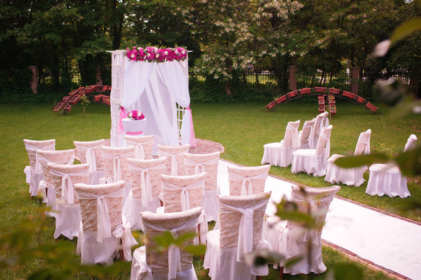 Элегантная розовая и белая свадебная арка со свежими цветами пионов, стоит на зеленой лужайке вместе с белыми дизайнерскими стульями
 - Фото, изображение
