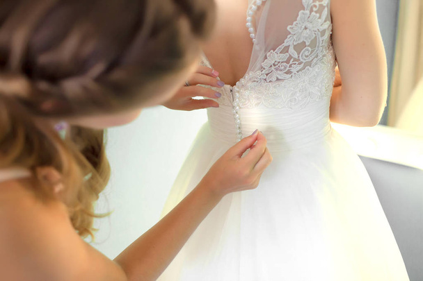 Bridesmaid helping slender bride lacing her wedding white dress, buttoning on delicate lace pattern with fluffy skirt on waist. Morning bridal preparation details newlyweds. Wedding day moments, wear - Valokuva, kuva