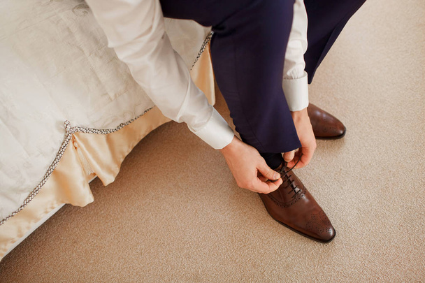 A man in a blue suit ties up shoelaces on brown leather shoes brogues on a wooden parquet background - Photo, Image