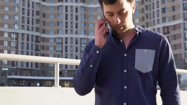 Subject businessman angry speaks on phone. Young caucasian brunette male in shirt. Small business worker. Profession. Sales manager. Realtor uses smartphone. Emotion anger and hatred against building - Filmati, video