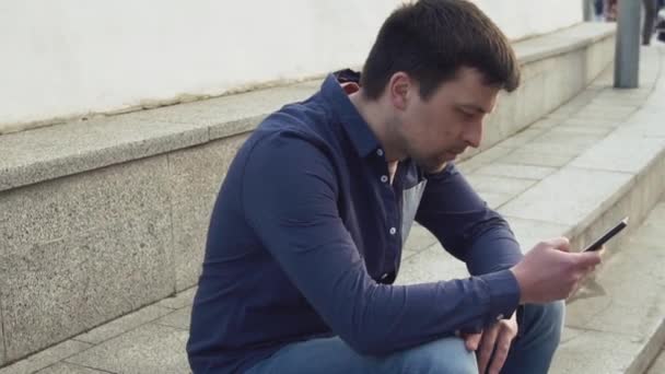 tematem jest człowiek i sieci społecznych. Kaukaski młody smutny mężczyzna brunetka w koszuli siedzi w parku w Letnec oczekiwania, spóźnienie jest ręką trzymać telefon komórkowy. Emocja urazy i zaburzenia - Materiał filmowy, wideo