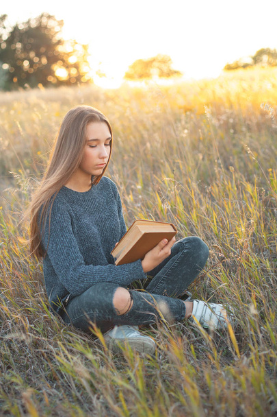 Όμορφη νεαρή γυναίκα κάθεται στο πεδίο και διαβάζοντας ένα βιβλίο στο ηλιοβασίλεμα - Φωτογραφία, εικόνα