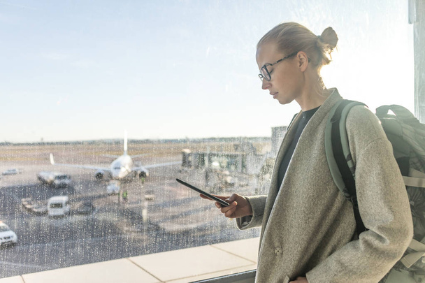 Satunnaisesti pukeutunut naispuolinen matkustaja lentokentällä katsomassa älypuhelimen laitteen edessä lentokentän portti ikkunat näkymät lentokenttien kiitotien - Valokuva, kuva
