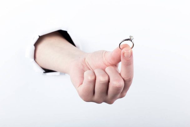 weibliche Hand aus einem Loch in Papier, hält einen Ehering, schließt eine Verlobung. isolieren auf weißem Hintergrund. - Foto, Bild