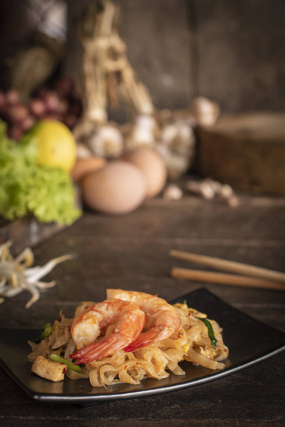 Thai fried noodles or pad thai with shrimp on black plate placed on the wood table there are chopsticks, eggs, lettuce, garlic, bean sprouts, shallot and cutting board placed back side. - Photo, Image