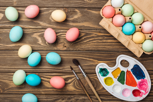top view of pastel easter eggs in cardboard box with color palette and paintbrushes on wooden table - Photo, Image