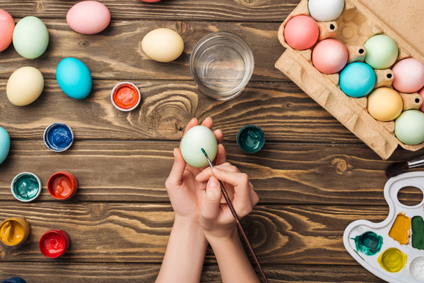 Vista parcial de niña decorando huevos de Pascua con pincel en mesa de madera con pinturas y paleta de colores
 - Foto, imagen