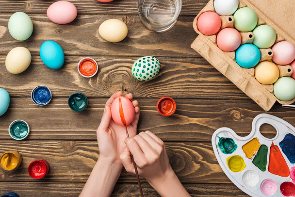 top view of woman decorating easter eggs with paintbrush at wooden table with paints and color palette - Фото, изображение