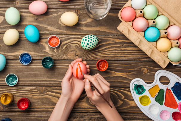 top view of woman decorating easter eggs with paintbrush at wooden table with paints and color palette - Foto, Imagen
