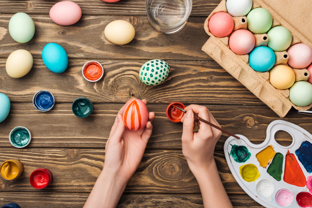 vista parcial de la mujer pintando huevos de Pascua en la mesa de madera con pinturas y paleta de colores
 - Foto, imagen