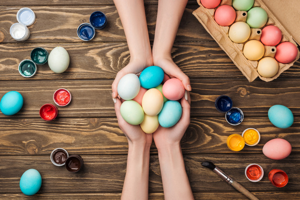 pohled shora ženy držící pastelové velikonoční vajíčka na dřevěný stůl s barvami - Fotografie, Obrázek