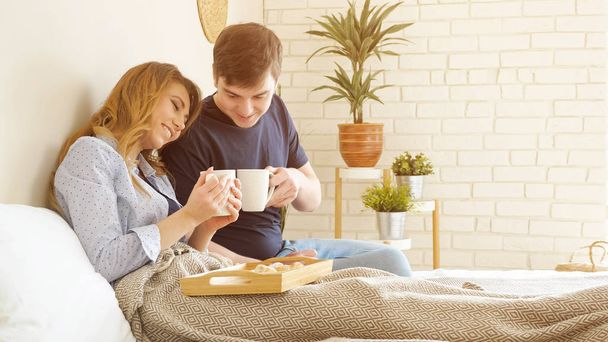 Young couple has breakfast in bed drinks tasty coffee miling in bedroom - Фото, изображение