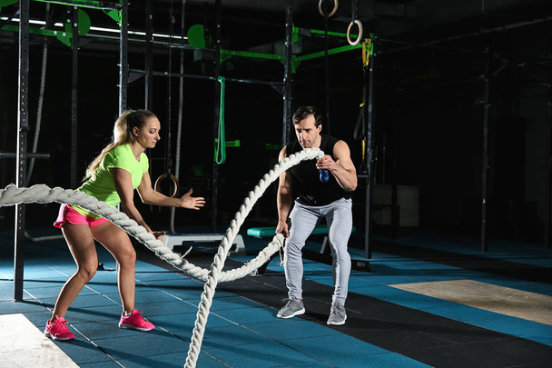 Young sporty man doing battle rope exercise under surveillance of trainer in gym - Foto, imagen