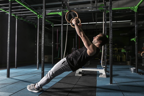 Young sporty man training with gymnastics rings in gym - Фото, изображение