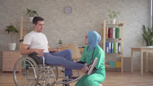 Muslim doctor in hijab examines the leg of a disabled person in a wheelchair - Séquence, vidéo