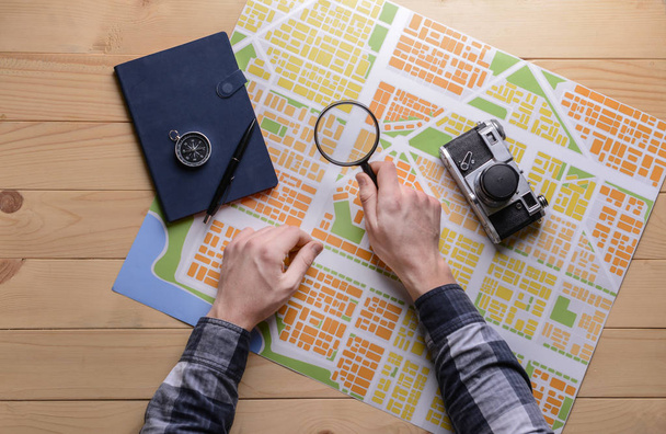 Mãos masculinas com mapa da cidade, lupa, bússola e câmera de fotos em mesa de madeira
 - Foto, Imagem