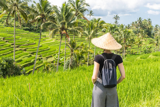 Rento muodikas valkoihoinen naisturisti yllään pieni reppu ja perinteinen aasialainen paddyhattu katselemassa kauniita vihreitä riisipeltoja ja terasseja Balilla saarella
 - Valokuva, kuva