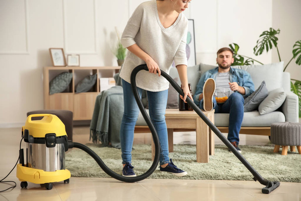 Vaimo puhdistaa lattiaa, kun hänen laiska miehensä katselee televisiota kotona
 - Valokuva, kuva