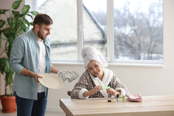 Carefree wife sitting at table while her indignant husband doing chores at home - Photo, Image