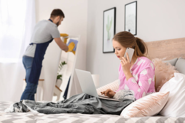 Πολυάσχολη γυναίκα που εργάζεται στο κρεβάτι, ενώ ο σύζυγός της να κάνουν δουλειές στο σπίτι - Φωτογραφία, εικόνα