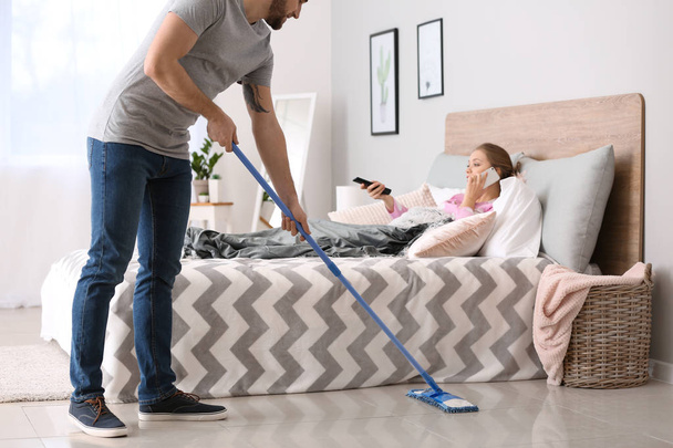 Househusband limpeza chão perto preguiçoso esposa deitada na cama
 - Foto, Imagem