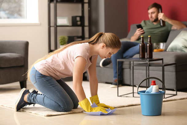 Vaimo puhdistaa lattiaa, kun hänen laiska miehensä lepää sohvalla kotona
 - Valokuva, kuva