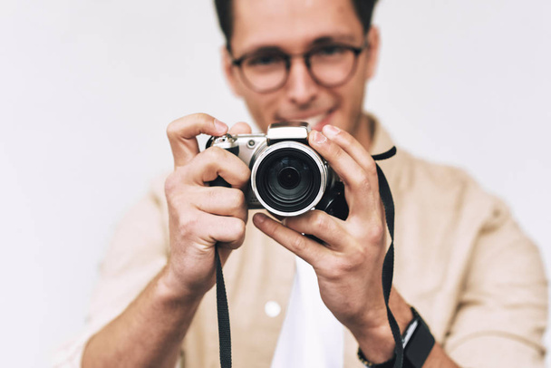 Image of young man smiling and taking photo on digital camera, posing over white studio background. Young male wearing casual outfit and trendy eyewear, blogging and looking to the camera display. - Foto, Imagem