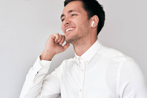 Young man smiling, having a call via wireless earphones with a colleague isolated on white wall. Caucasian businessman wearing white shirt, using wireless earbuds for communication. Technology, people - Photo, Image