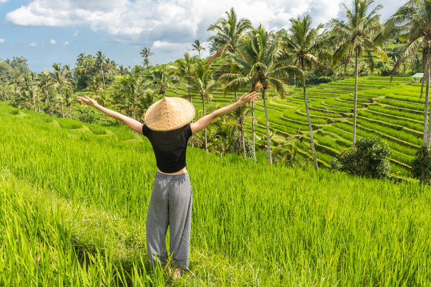 Relaxed fashionable female traveler wearing small backpack and traditional asian paddy hat, arms rised to sky, enjoying pure nature at beautiful green rice fields and terraces on Bali island - Zdjęcie, obraz